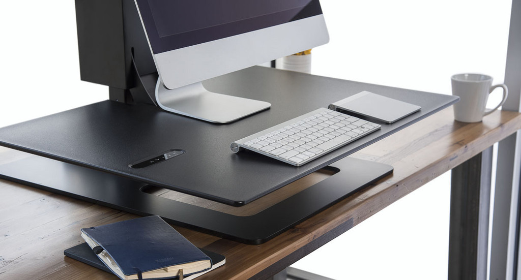 UPLIFT E7 UDA115 electric desk converter shown with a Black Eco work surface on a host desk; the pictured mount arrangement is not a verified match for the selected single-mount option