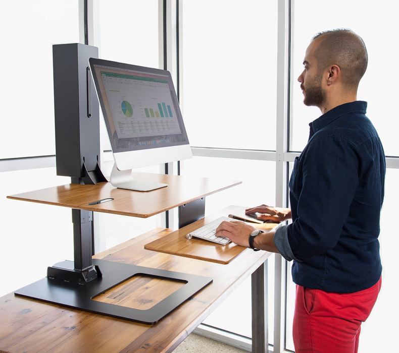 UPLIFT E7 electric desk converter on an existing desk, with a raised wood-tone work surface, dark base, lower keyboard tray and one mounted monitor