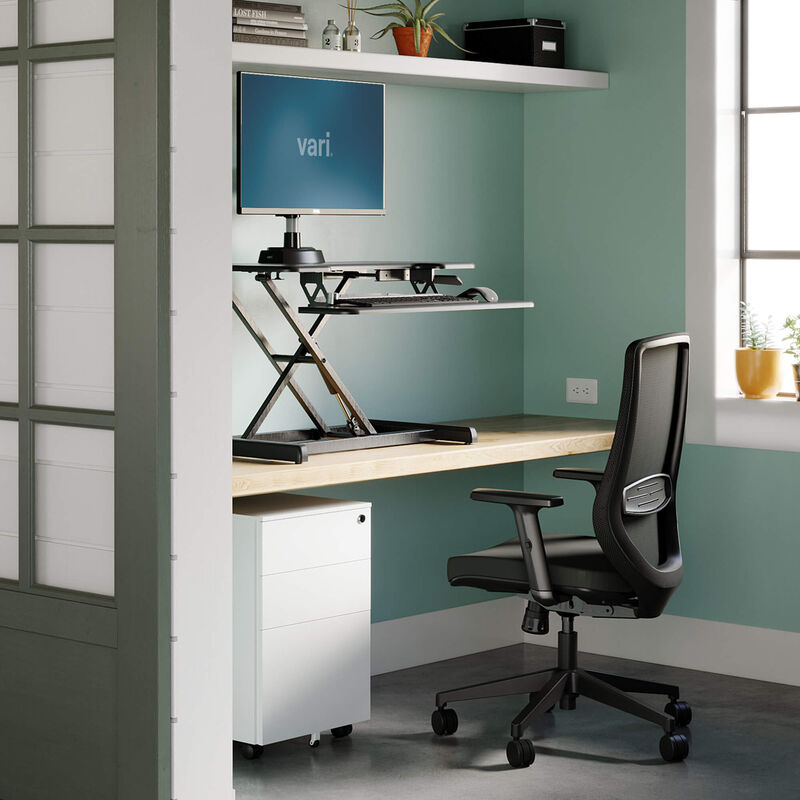 VariDesk Essential Vertical Lift 30 in black, raised on its linked product-page image; separate host desk and staged equipment are context.